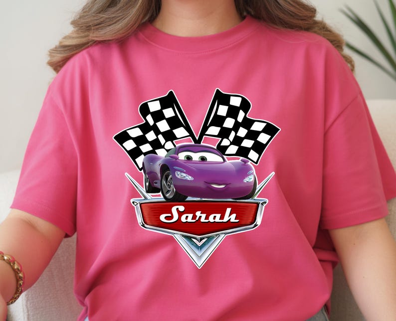 May include: A magenta t-shirt featuring a purple cartoon car with two checkered flags. The car has a smiling face and is set against a red banner with the name "Sarah".