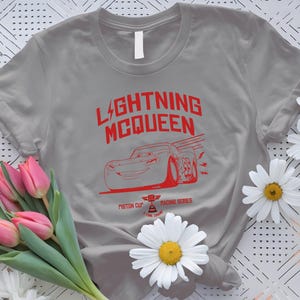 May include: A gray t-shirt featuring the text "LIGHTNING MCQUEEN" in red, along with a red graphic of a race car. The shirt also includes the text "Piston Cup Racing Series".