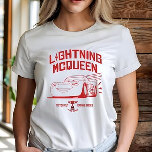 May include: White t-shirt featuring a red graphic of Lightning McQueen from the movie Cars. The design includes the text "LIGHTNING MCQUEEN" above an illustration of the race car, along with "PISTON CUP" and "RACING SERIES".