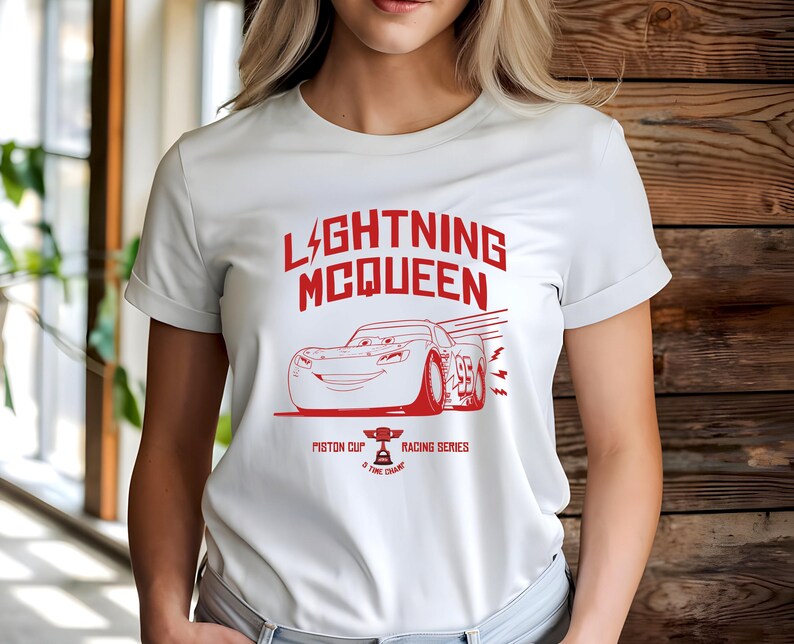 May include: White t-shirt featuring a red graphic of Lightning McQueen from the movie Cars. The design includes the text "LIGHTNING MCQUEEN" above an illustration of the race car, along with "PISTON CUP" and "RACING SERIES".