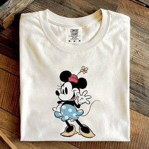 May include: A cream-colored short-sleeve t-shirt featuring a graphic of Minnie Mouse wearing a red hat, a blue skirt with white polka dots, and tan shoes. The character has a small flower on her hat. The shirt has a Comfort Colors brand label.