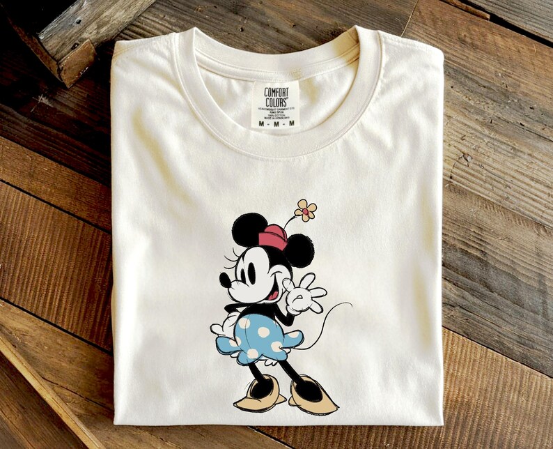 May include: A cream-colored short-sleeve t-shirt featuring a graphic of Minnie Mouse wearing a red hat, a blue skirt with white polka dots, and tan shoes. The character has a small flower on her hat. The shirt has a Comfort Colors brand label.