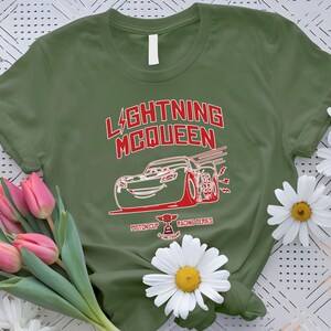 May include: Olive green t-shirt featuring a red and white graphic of Lightning McQueen from the movie Cars. The design includes the text "LIGHTNING McQUEEN" and "Piston Cup Racing Series". The shirt is decorated with pink tulips and white daisies.