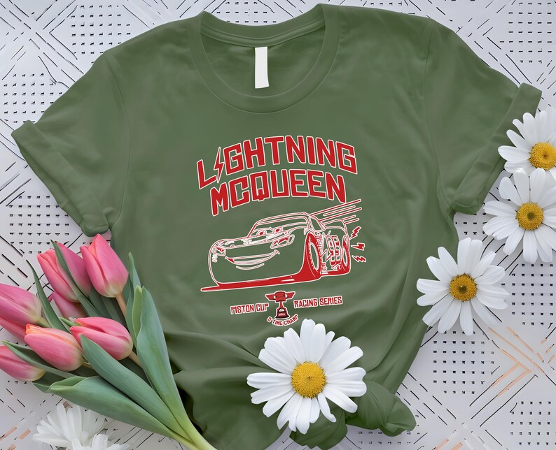 May include: Olive green t-shirt featuring a red and white graphic of Lightning McQueen from the movie Cars. The design includes the text "LIGHTNING McQUEEN" and "Piston Cup Racing Series". The shirt is decorated with pink tulips and white daisies.