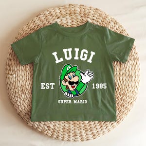 May include: Green t-shirt featuring a graphic of Luigi from Super Mario. The design includes the name "LUIGI" in white block letters above an image of Luigi, with the text "EST 1985" and "SUPER MARIO" below. The shirt is displayed on a woven surface.