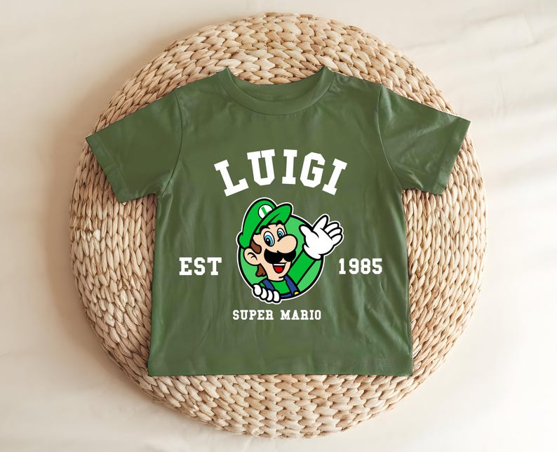 May include: Green t-shirt featuring a graphic of Luigi from Super Mario. The design includes the name "LUIGI" in white block letters above an image of Luigi, with the text "EST 1985" and "SUPER MARIO" below. The shirt is displayed on a woven surface.