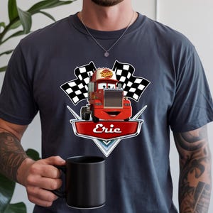 May include: Dark gray t-shirt featuring a red cartoon truck with the name "Eric" on a shield. The truck has a smiling face and is flanked by two checkered flags. The word "Rusteze" is on the truck's roof.