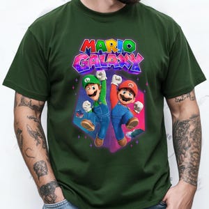 Puede incluir: Camiseta verde oscuro con la inscripci&oacute;n "MARIO GALAXY" en letras 3D coloridas. El dise&ntilde;o incluye ilustraciones de Mario y Luigi, con los brazos levantados en se&ntilde;al de celebraci&oacute;n. Los personajes se encuentran sobre un fondo geom&eacute;trico vibrante.