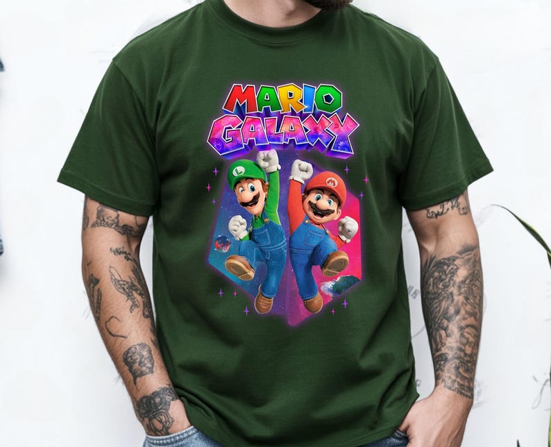 Puede incluir: Camiseta verde oscuro con la inscripci&oacute;n "MARIO GALAXY" en letras 3D coloridas. El dise&ntilde;o incluye ilustraciones de Mario y Luigi, con los brazos levantados en se&ntilde;al de celebraci&oacute;n. Los personajes se encuentran sobre un fondo geom&eacute;trico vibrante.
