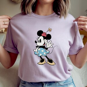 May include: A light purple short-sleeved t-shirt featuring a graphic of Minnie Mouse. The character is depicted with a red hat, a blue skirt with white polka dots, and yellow shoes, while holding a small yellow flower above her head.
