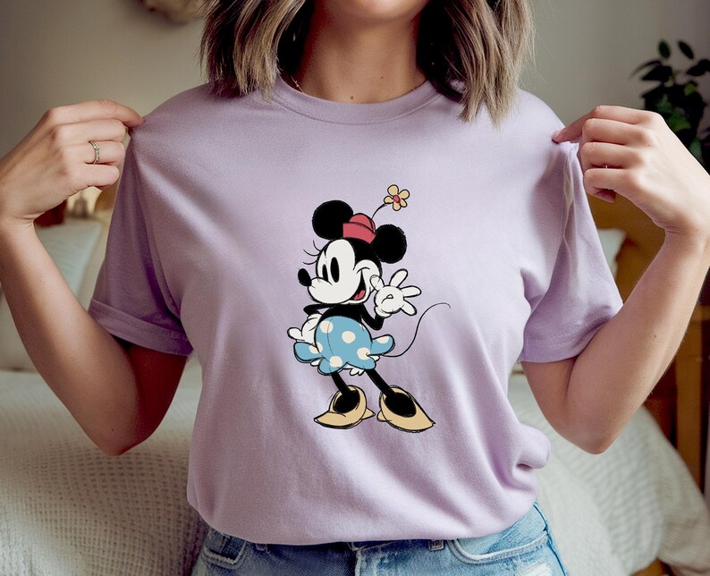 May include: A light purple short-sleeved t-shirt featuring a graphic of Minnie Mouse. The character is depicted with a red hat, a blue skirt with white polka dots, and yellow shoes, while holding a small yellow flower above her head.
