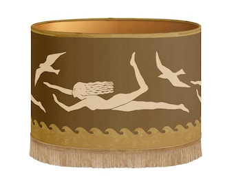 May include: A cylindrical lampshade with a brown background and a beige design. The design features a woman swimming, birds, and waves. The lampshade has a gold interior and a fringe at the bottom.