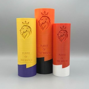 Peut inclure: Trois trophées cylindriques jaunes, oranges et rouges, chacun avec un motif de tête de lion et de couronne. Le texte "PLAYER OF THE MATCH" est imprimé sur chaque trophée. Les bases sont violettes, noires et blanches.