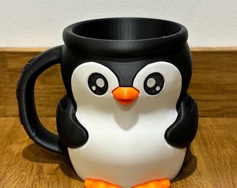 Puede incluir: Taza impresa en 3D con forma de pingüino. La taza presenta un borde, asa y detalles en negro, un cuerpo blanco y un pico y patas naranjas. Los ojos del pingüino son grandes y detallados, creando un diseño divertido.
