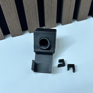 Puede incluir: Un juego de accesorios de c&aacute;mara negros impresos en 3D. El juego incluye un componente principal cuadrado con una abertura circular, una base rectangular y dos peque&ntilde;as piezas en forma de L. Los art&iacute;culos est&aacute;n sobre una superficie blanca.