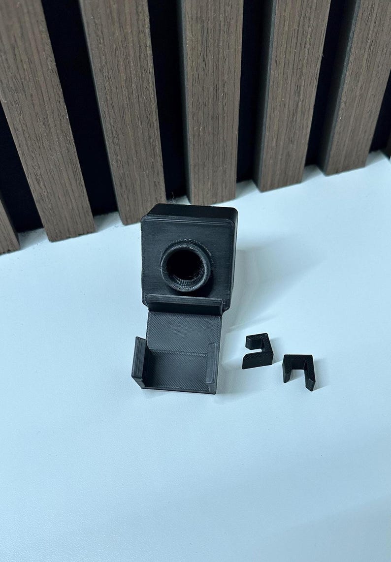 Puede incluir: Un juego de accesorios de c&aacute;mara negros impresos en 3D. El juego incluye un componente principal cuadrado con una abertura circular, una base rectangular y dos peque&ntilde;as piezas en forma de L. Los art&iacute;culos est&aacute;n sobre una superficie blanca.