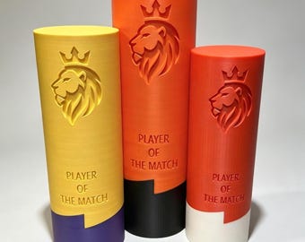 Puede incluir: Tres trofeos cilíndricos de diferentes alturas en amarillo, naranja y rojo. Cada trofeo presenta una cabeza de león en relieve con una corona sobre el texto "PLAYER OF THE MATCH". Las bases son de color morado, negro y blanco respectivamente.