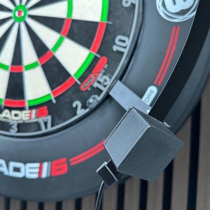 Może przedstawiać: Zbliżenie na tarczę do darta z czarno-białym wzorem tarczy, z czerwonymi i zielonymi pierścieniami akcentującymi. Na tablicy znajduje się napis "BLADE 6" w kolorze czerwonym i białym. Do tablicy przymocowane jest czarne, kwadratowe akcesorium.