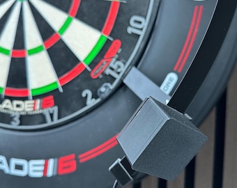 Puede incluir: Primer plano de un tablero de dardos con un diseño de diana en blanco y negro, con anillos de acento rojos y verdes. El tablero tiene el texto "BLADE 6" en rojo y blanco. Un accesorio negro de forma cuadrada está unido al tablero.