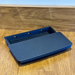Può includere: Una scatola rettangolare blu scuro con un coperchio strutturato, parzialmente aperto. La scatola ha tre fori circolari all'interno. La scatola è su una superficie di legno.