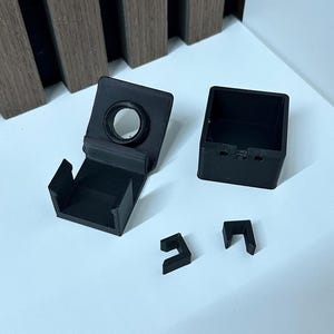 Puede incluir: Componentes negros impresos en 3D, que incluyen una caja cuadrada, un soporte con una abertura circular y dos piezas en forma de L. Los objetos est&aacute;n dispuestos sobre una superficie blanca, con un panel de madera en el fondo.