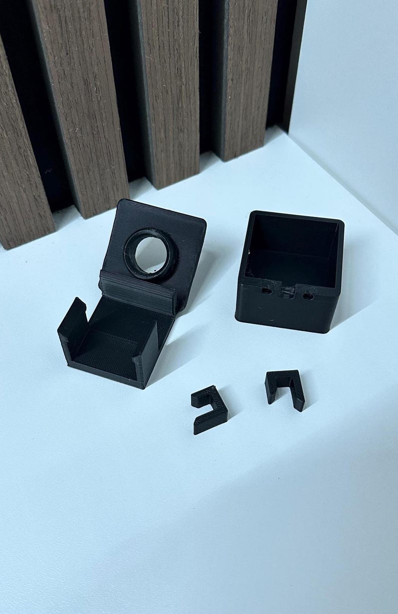 Puede incluir: Componentes negros impresos en 3D, que incluyen una caja cuadrada, un soporte con una abertura circular y dos piezas en forma de L. Los objetos est&aacute;n dispuestos sobre una superficie blanca, con un panel de madera en el fondo.