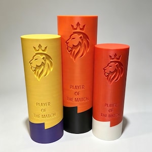 May include: Three cylindrical trophies of varying heights, each featuring an embossed lion head with a crown and the text PLAYER OF THE MATCH. The yellow trophy has a purple base, the orange trophy has a black base, and the red trophy has a white base.