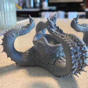May include: A gray, 3D-printed octopus sculpture with multiple tentacles. The tentacles are curved and textured with small, pointed protrusions. The sculpture is resting on a light-colored countertop, with blurred kitchen items in the background.