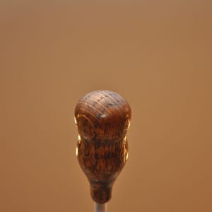 May include: A close-up of a wooden bottle stopper with a rounded top and a textured, dark brown finish. The stopper has a bulbous shape and a slender stem, set against a warm, neutral background. The wood grain is visible.
