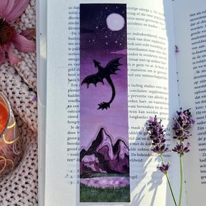 Op de afbeelding: Een fantasy-thema bladwijzer met een draak silhouet tegen een paarse nachthemel met een volle maan. De bladwijzer staat in een open boek, met een kaars en een bloem.