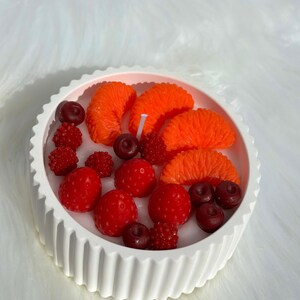 May include: A white, fluted-edge candle holder filled with a candle and wax fruit. The wax fruit includes bright orange mandarin orange slices, red strawberries, cherries, and raspberries. A white candle wick is visible in the center.