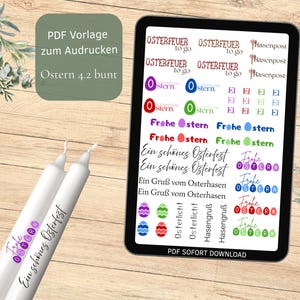 Op de afbeelding: Een digitale tablet toont Pasen-thema graphics en tekst, waaronder "Frohe Ostern" en "Ein schönes Osterfest". Twee witte kaarsen met paarse tekst en een bloemenontwerp staan op de voorgrond. De achtergrond is een houten oppervlak.