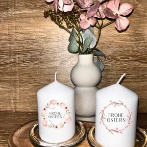 Könnte beinhalten: Zwei weiße Stumpenkerzen mit dem Text "FROHE OSTERN" und einem floralen Kranzdesign. Eine Vase mit rosa Blumen steht hinter den Kerzen auf einem Holztablett. Die Kerzen stehen auf Glasschalen.