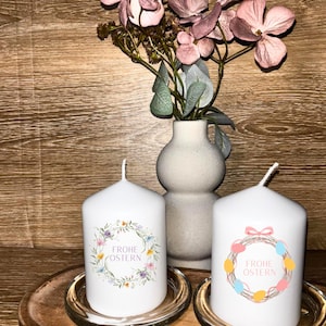 Könnte beinhalten: Zwei weiße Stumpenkerzen mit floralen Kranzdesigns und dem Text "FROHE OSTERN" stehen auf Glasschalen. Eine Vase mit rosa Blumen und ein Holztablett befinden sich im Hintergrund. Die Kerzen sind ein dekorativer Artikel.