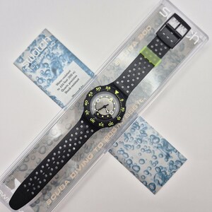 May include: A black Swatch Scuba 200 watch with a perforated strap and a black buckle. The watch face is black with white and neon green accents. The watch is water-resistant to 200 m. The watch is in a clear plastic case with a blue and white card.