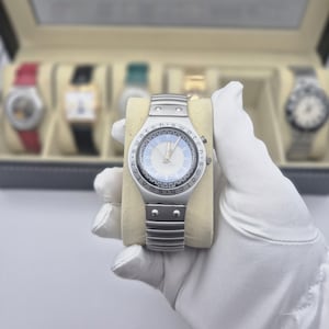 May include: A silver-toned wristwatch with a light blue face and silver hands. The watch has a metal band and is held on a beige pillow. Other watches are visible in the background.