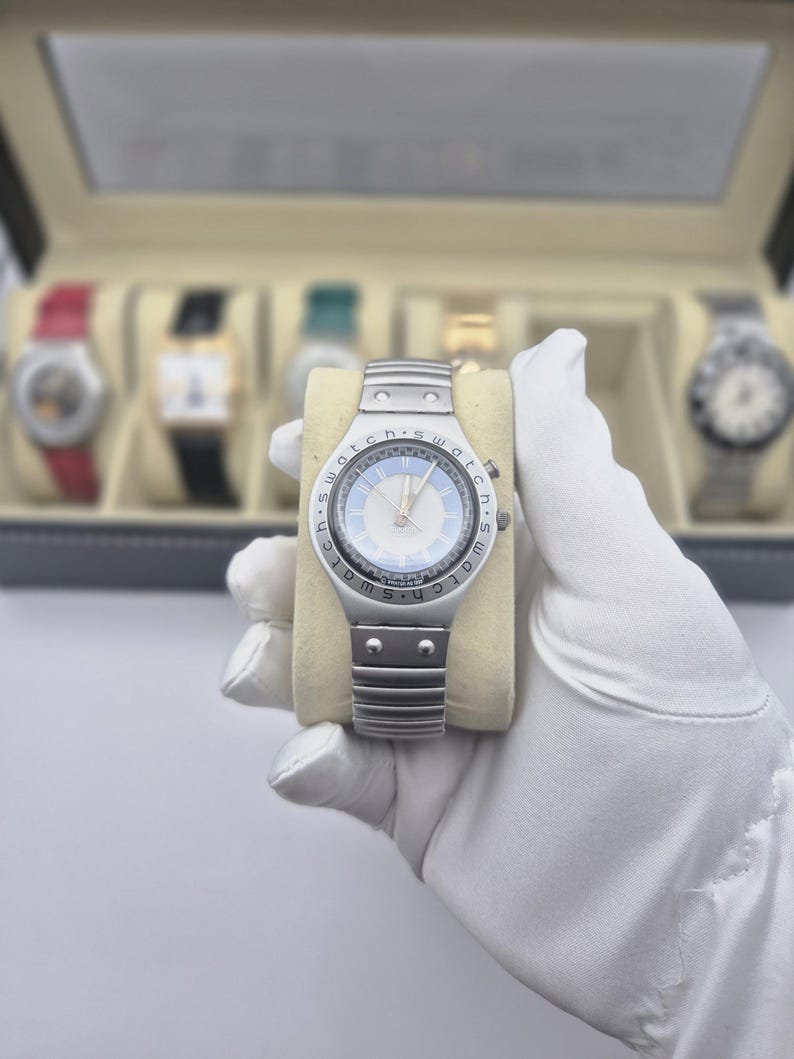 May include: A silver-toned wristwatch with a light blue face and silver hands. The watch has a metal band and is held on a beige pillow. Other watches are visible in the background.