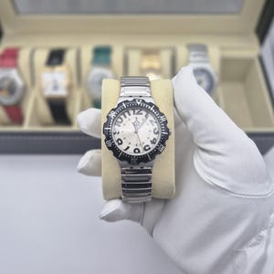 May include: A silver-toned wristwatch with a black bezel and a white face. The watch has a silver band and is displayed on a beige cushion. The watch face features black numerals and the word "JACOBS". Other watches are visible in the background.