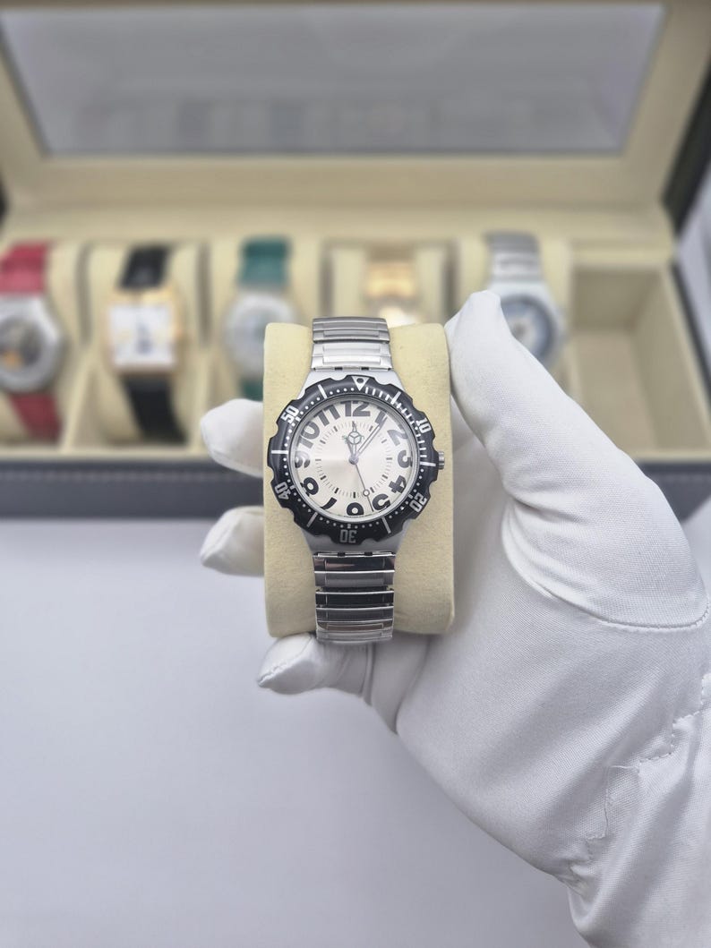 May include: A silver-toned wristwatch with a black bezel and a white face. The watch has a silver band and is displayed on a beige cushion. The watch face features black numerals and the word "JACOBS". Other watches are visible in the background.