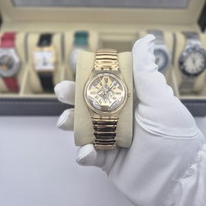 May include: A gold-toned wristwatch with a skeleton dial and a matching gold-toned band. The watch features Roman numeral hour markers and is displayed on a beige cushion. Other watches are visible in the background.