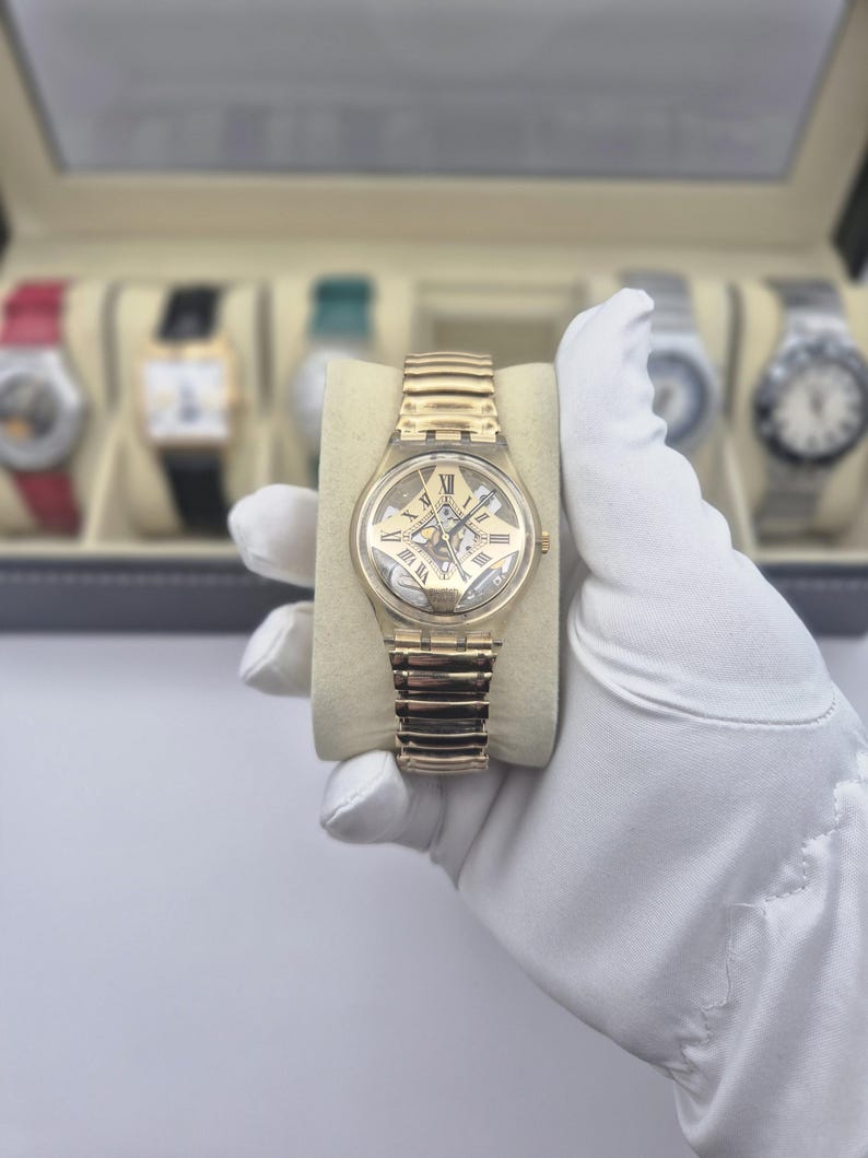 May include: A gold-toned wristwatch with a skeleton dial and a matching gold-toned band. The watch features Roman numeral hour markers and is displayed on a beige cushion. Other watches are visible in the background.