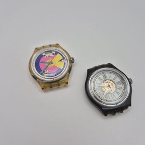 Op de afbeelding: Twee polshorloges. Eén heeft een transparante gele kast met een kleurrijke wijzerplaat met roze, gele en blauwe segmenten. Het andere horloge heeft een zwarte kast, een witte wijzerplaat met Romeinse cijfers en een datumweergave.