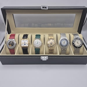 May include: A black watch display case with a clear glass top, showcasing six assorted wristwatches. The watches feature various band colors, including red, black, green, and silver. The case is lined with a light beige material.
