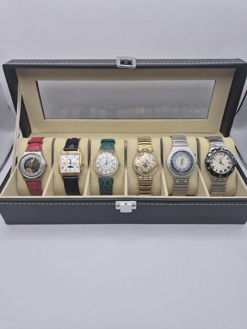 May include: A black watch display case with a clear glass top, showcasing six assorted wristwatches. The watches feature various band colors, including red, black, green, and silver. The case is lined with a light beige material.