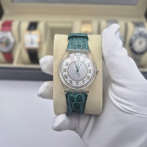 May include: A clear Swatch watch with a silver dial and Roman numeral hour markers. The watch has a teal leather band with a crocodile pattern. The watch is held on a beige cushion.