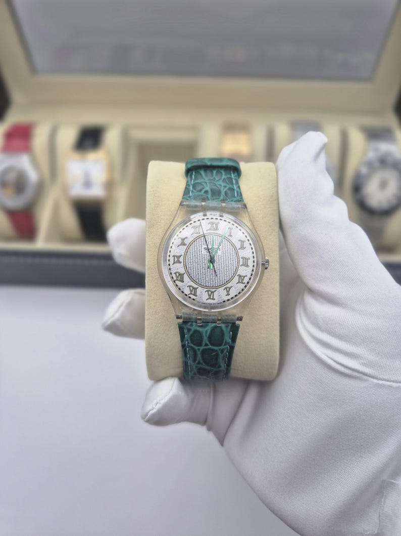 May include: A clear Swatch watch with a silver dial and Roman numeral hour markers. The watch has a teal leather band with a crocodile pattern. The watch is held on a beige cushion.