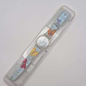 May include: A Swatch watch in its original packaging. The watch features a light blue strap adorned with colorful butterfly and dragonfly illustrations. The watch face is silver, and the strap is in a clear plastic case.
