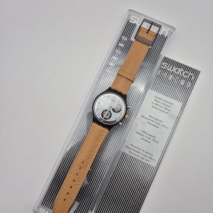 May include: A white ceramic mug with a black handle and the text "But First, Coffee" in a bold, black font. The mug is set against a blurred background, suggesting a cozy setting.