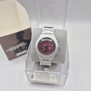 May include: White wristwatch with a red face, displayed in a clear plastic case. The watch has a white band and a round face with multiple dials. The watch is presented on a gray cushion, with a box in the background.