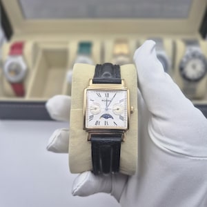 May include: A gold-toned rectangular Fossil watch with a white face, black leather band, and Roman numeral hour markers. The watch features a moon phase indicator and is displayed on a beige cushion.
